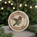 A round ornament featuring a hummingbird with a red head and brown body, surrounded by greenery and a string of lights in the background.