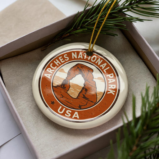 A circular ornament with the text "Arches National Park USA" and an illustration of a rock formation, placed in a box with pine branches.