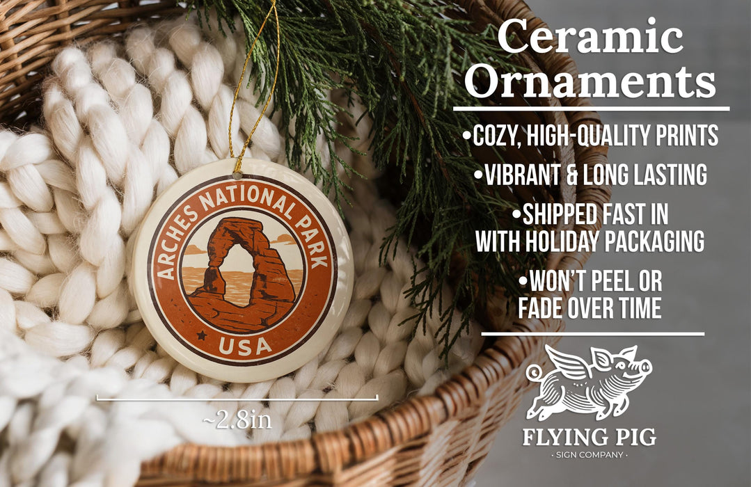 Ceramic ornaments, featuring a round ornament with the text "Arches National Park USA" and an illustration of the Arches National Park in the United States. The ornament is hanging from a wicker basket, and there is a pine branch next to it.