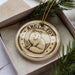 A circular ornament with a drawing of an Arctic fox, surrounded by stars, is displayed in a box with pine branches.
