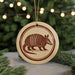 A round ornament featuring an armadillo design is displayed in front of a Christmas tree with twinkling lights.