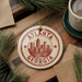 A circular ornament with the text "ATLANTA GEORGIA" and a graphic of a city skyline is displayed on a wooden surface, surrounded by pine branches and a cup of coffee.