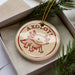 A round ornament with the word "AXOLOTL" printed on it, featuring an illustration of an axolotl, a type of salamander. The ornament is placed inside a box with pine branches, creating a festive and nature-inspired display.