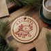 A round ornament featuring an axolotl, a type of salamander, is displayed on a wooden surface alongside a cup of coffee and some pine branches.