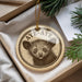 A round ornament with a picture of a cute animal, possibly a cat or a dog, and the text "Aye-aye" printed on it, placed in a box with pine branches.