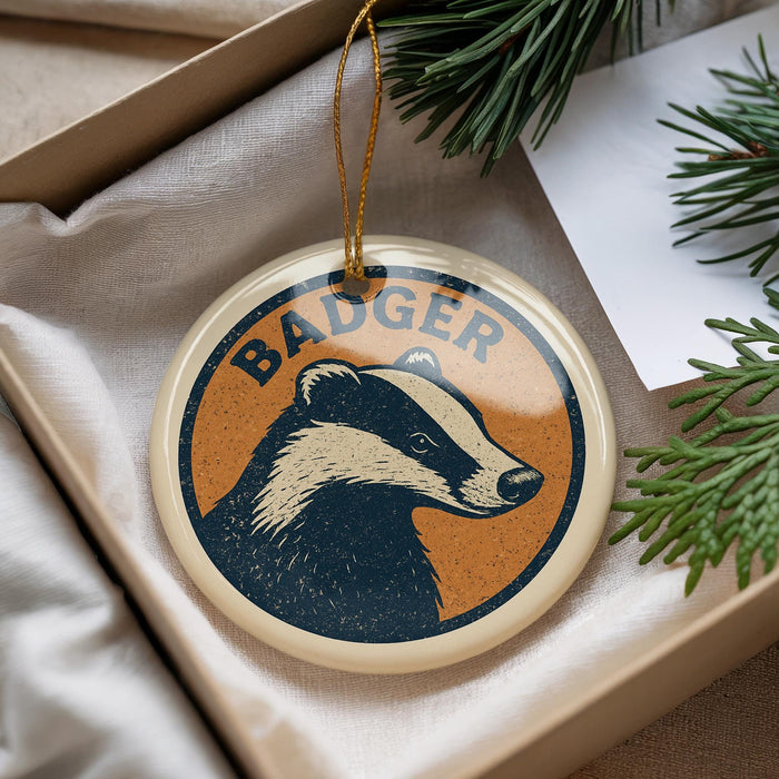 A circular ornament with a badger's face design, featuring the word "BADGER" written on it, is displayed in a box with pine branches.