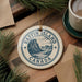 A circular ornament with the text "Baffin Island Canada" printed on it, surrounded by pine branches and a cup of coffee on a wooden surface.