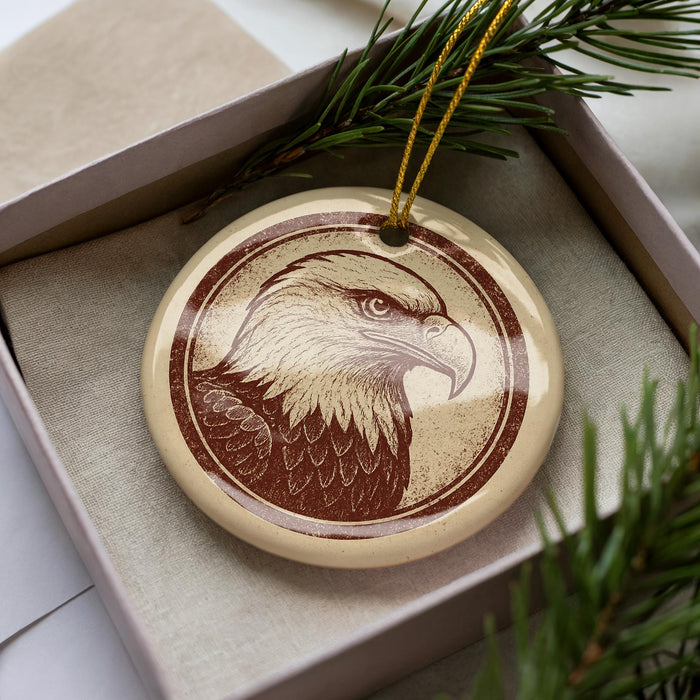 A round ornament featuring a detailed illustration of an eagle is displayed in a box, surrounded by pine branches.