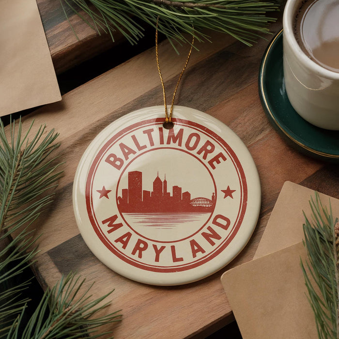 A round ornament with the text "BALTIMORE MARYLAND" printed on it, featuring a silhouette of a city skyline. The ornament is hanging from a string, and there are pine branches and a cup of coffee visible in the background.