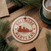 A round ornament with the text "BALTIMORE MARYLAND" printed on it, featuring a silhouette of a city skyline. The ornament is hanging from a string, and there are pine branches and a cup of coffee visible in the background.