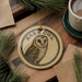 A round ornament featuring a barn owl design, with the text "Barn Owl" prominently displayed. The ornament is placed on a wooden surface, surrounded by pine branches and a cup of coffee.
