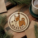 A circular ornament featuring a brown and white dog, with the name "Basesenji" written on it, is displayed on a wooden surface alongside a cup of coffee and some pine branches.