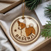 A circular ornament featuring a brown and white dog, with the name "BASENJI" written on it, is displayed in a box with pine branches.