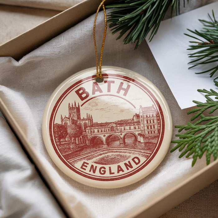 A circular ornament with a red and white design, featuring a castle and a bridge, is displayed in a box with pine branches.