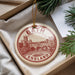 A circular ornament with a red and white design, featuring a castle and a bridge, is displayed in a box with pine branches.