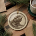 a ceramic ornament with a detailed illustration of a bearded dragon, placed on a wooden surface alongside a cup of coffee and some pine branches.