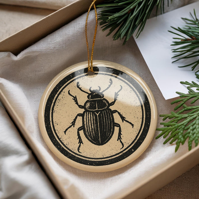 A decorative ornament featuring a black and white illustration of a beetle is displayed in a box, surrounded by pine branches.