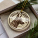 A circular ornament with a dog's head and the text "Belgian Malinois" printed on it, placed inside a box with pine branches.