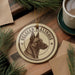 A circular ornament featuring a dog's head and the text "Belgian Malinois" is displayed on a wooden surface, surrounded by pine branches and a cup of coffee.