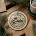 A circular ornament with the text "BEQUIA SAINT VINCENT AND THE GREENVADORINES" printed on it, surrounded by pine branches and a cup of coffee on a wooden surface.