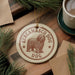 A circular ornament featuring a shaggy dog, likely a Berganco dog, is displayed on a wooden surface alongside a cup of coffee and some pine branches.