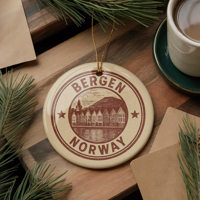 A round ornament with the text "BERGEN NORWAY" and a graphic of a town is displayed on a wooden surface, surrounded by pine branches and a cup of coffee.
