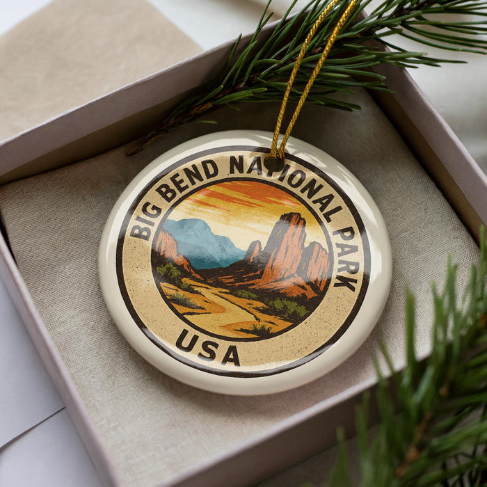 A decorative ornament featuring the Big Bend National Park logo, which is a circular emblem with a mountain landscape and the text "Big Bend National Park USA" printed on it. The ornament is placed inside a box, surrounded by pine branches.