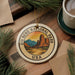 A decorative ornament featuring the Big Bend National Park logo, surrounded by pine branches and a cup of coffee on a wooden surface.