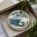 A circular ornament with a picture of a bear and the word "Binturong" on it, placed in a box with pine branches.