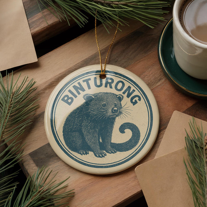 A round ornament featuring a cartoon image of a raccoon, with the word "Binturong" written on it, is displayed on a wooden surface alongside a cup of coffee and some pine branches.