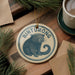 A round ornament featuring a cartoon image of a raccoon, with the word "Binturong" written on it, is displayed on a wooden surface alongside a cup of coffee and some pine branches.