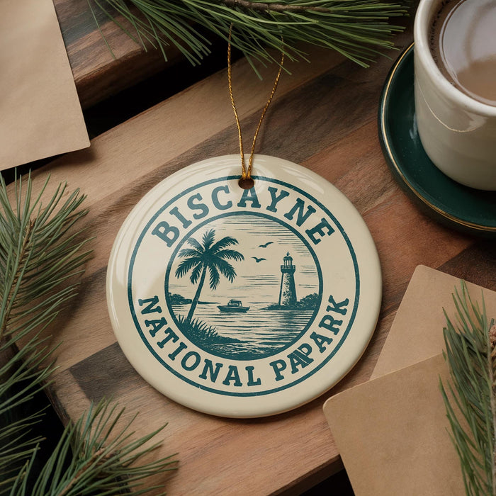 A ceramic ornament with the text "Biscayne National Pap Park" and a lighthouse image, placed on a wooden surface with pine branches and a cup of coffee nearby.