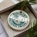 A white ceramic ornament with the text "BISCAYNE NATIONAL PAPARK" printed on it, placed in a box with pine branches.