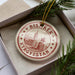 A circular ornament with the text "BISKECK" and "Kyrgyzstan" printed on it, surrounded by a border of stars. The ornament is placed inside a box with pine branches.