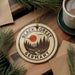 A circular ornament with the text "BLACK FOREST GERMANY" and a landscape illustration, hanging on a wooden surface surrounded by pine branches and a cup of coffee.
