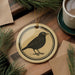 a round ornament with a black bird illustration on it, placed on a wooden surface next to a cup of coffee and some pine branches.