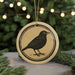 A round ornament featuring a black bird is displayed in front of a blurred Christmas tree with twinkling lights.