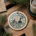 A decorative ornament with the text "BLOCK ISLAND RHODE ISLAND, USA" on it, surrounded by pine branches and a cup of coffee on a wooden surface.