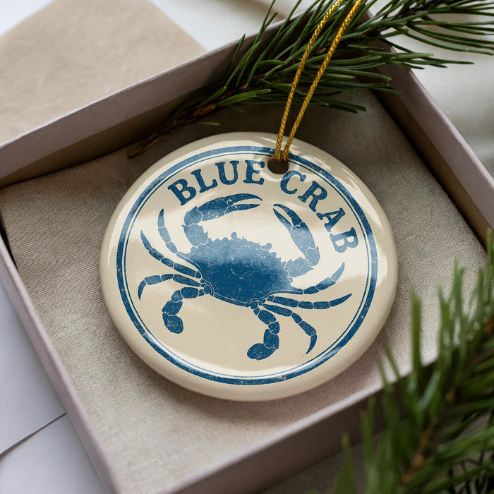 A round ornament with a blue crab design, placed inside a box with pine branches.