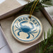 A round ornament with a blue crab design, placed inside a box with pine branches.