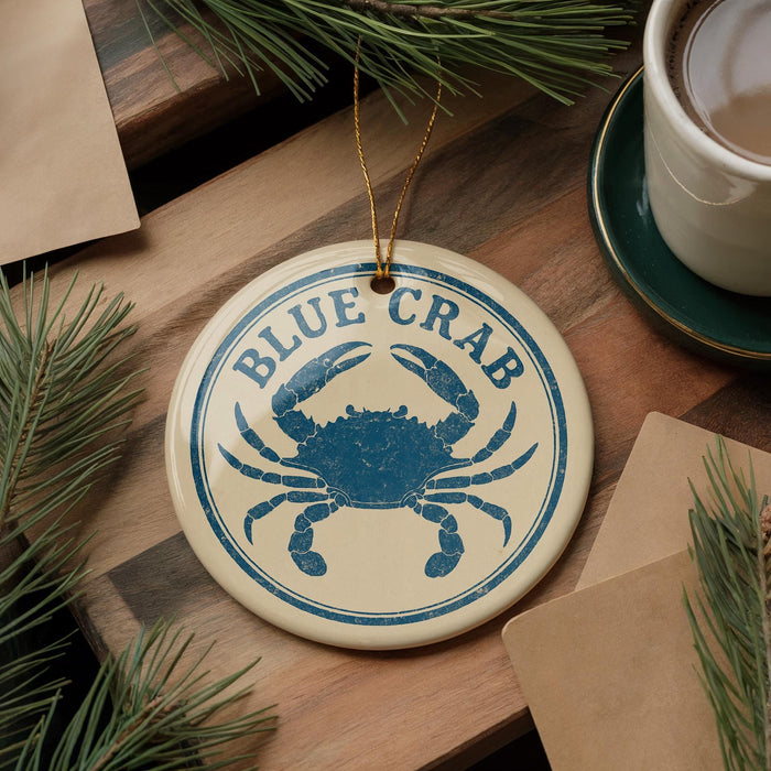 A blue crab ornament is displayed on a wooden surface, surrounded by pine branches and a cup of coffee.