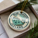 A circular ornament with the text "BLUE LAGOON ICELAND" and an illustration of a scenic landscape, including a body of water and mountains, is displayed in a box with pine branches.