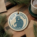 A blue and white ornament featuring a monkey is displayed on a wooden surface, surrounded by pine branches and a cup of coffee.