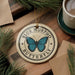 A circular ornament with a blue butterfly design, featuring the text "BLUE MORPHO BUTTERFLY" on it, is displayed on a wooden surface alongside a cup of coffee and some pine branches.