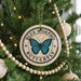 A circular ornament with a blue butterfly design hangs from a Christmas tree, surrounded by other ornaments and lights.