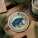 A blue poison dart frog ornament is displayed on a wooden surface, surrounded by pine branches and a cup of coffee.