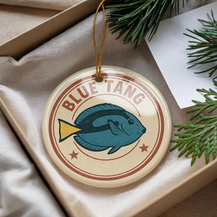 A round ornament with a blue fish design and the text "BLUE TANG" is displayed in a box, surrounded by pine branches.