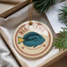 A round ornament with a blue fish design and the text "BLUE TANG" is displayed in a box, surrounded by pine branches.