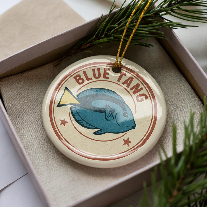 A round ornament with a blue fish design and the text "BLUE TANG" is displayed in a box, surrounded by pine branches.