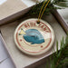 A round ornament with a blue fish design and the text "BLUE TANG" is displayed in a box, surrounded by pine branches.